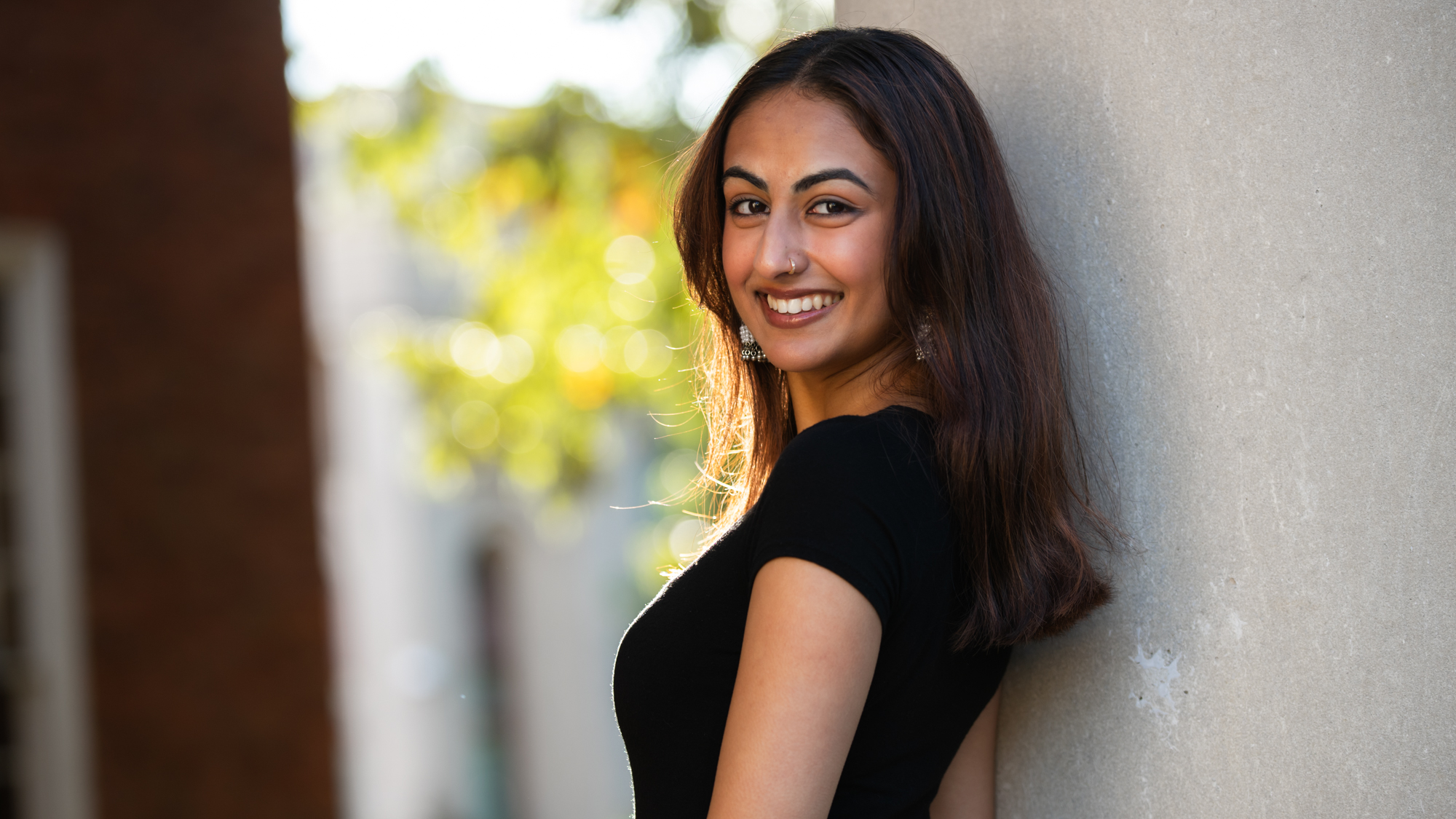 Keya at UNC in a black dress and silver earrings, leaning against a pillar and smiling